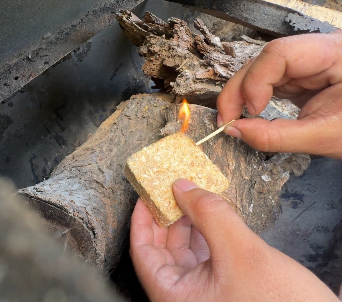A lit match touching the corner of a One Strike fire starter held over firewood