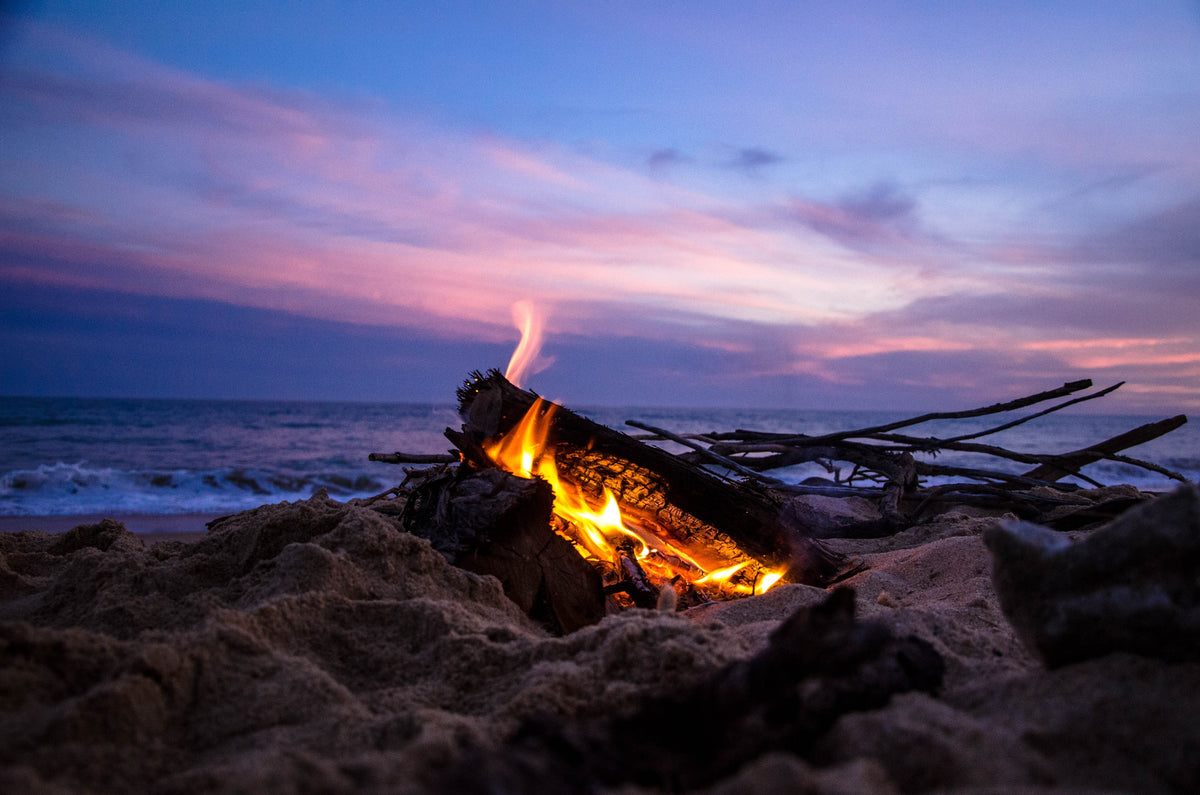 A driftwood campfire burning on a beach at dusk under a pink and violet sky